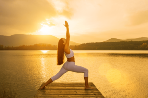 Femme effectuant la pose de yoga du Virabhadrasana 1 ou posture du guerrier au coucher du soleil sur un ponton avec la vue sur la mer.