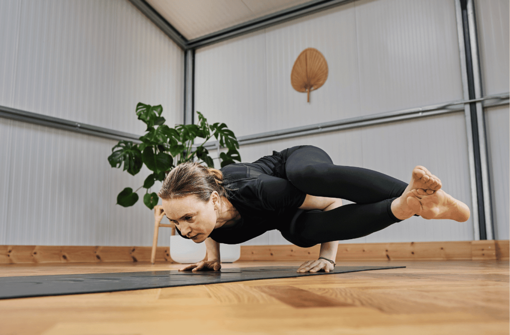 Femme pratiquant la pose de yoga du corbeau latéral, corps parallèle au sol et jambes croisées, dans une pièce avec une plante en arrière-plan.