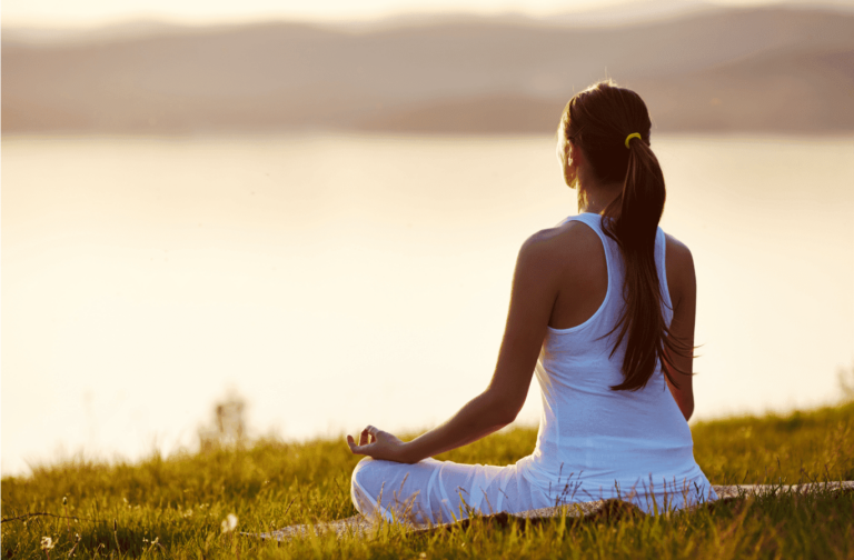 Femme assise en méditation en plein air, vue de dos, au coucher du soleil avec une vue sur le lac et les montagnes en arrière-plan.